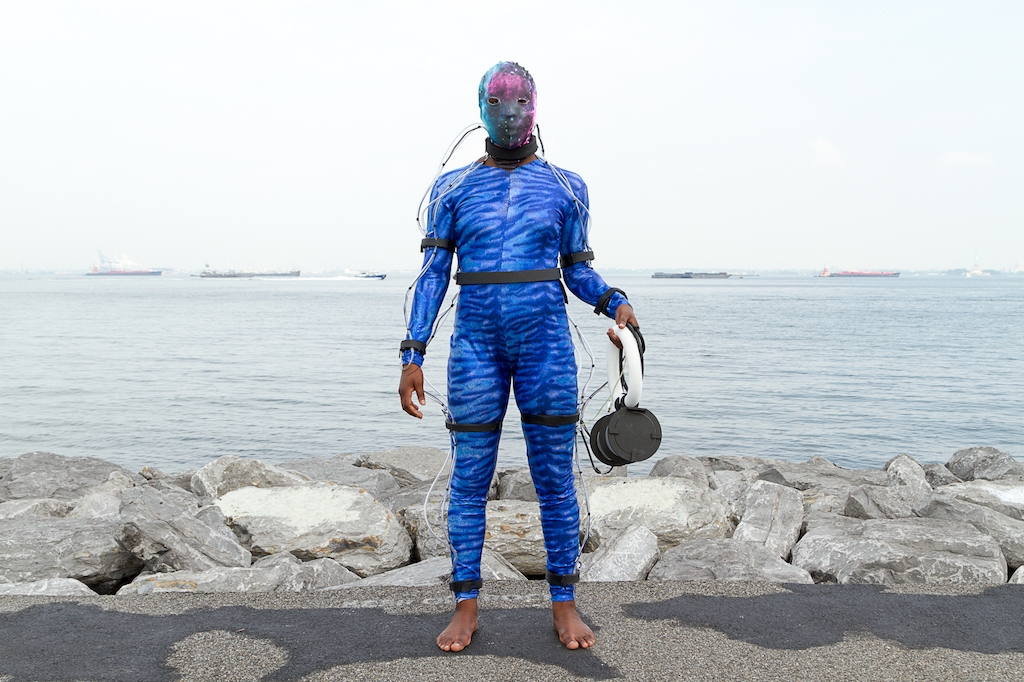 Person stands at waters edge in futuristic wet suit with wires attached to a facemask