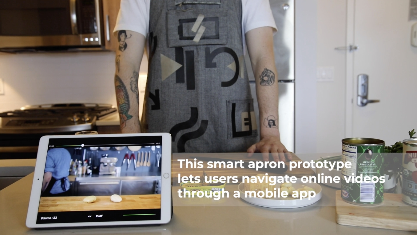 Someone wearing a smart apron stands in front of a cutting board, text on the screen reads "this smart apron prototype lets users navigate online videos through a mobile app"