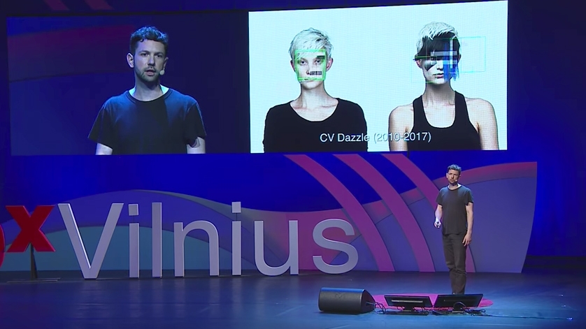 A person on stage giving a talk with a projection of 2 people overlaid with facial recognition graphics