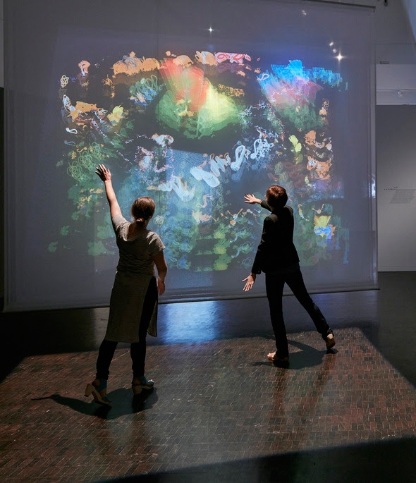 2 people standing and interacting with a projection that is displaying colorful patterns