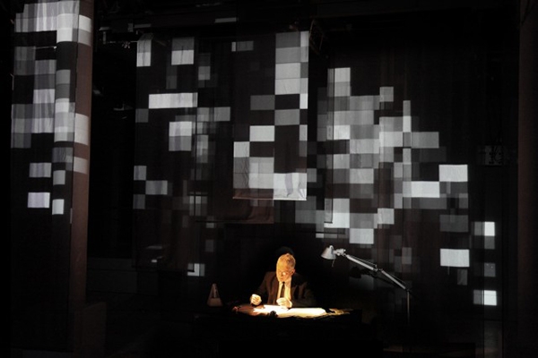A wide shot of a man at a desk illuminated by a desk lamp with pixelated rectangles projected behind him