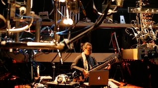 a wideshot of a person playing an eclectic guitar surrounded by other experimentally looking instruments