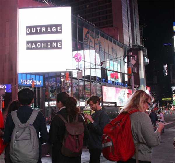 Times Square with the logo of Outrage Machine on display, with a few people looking at their cell phones