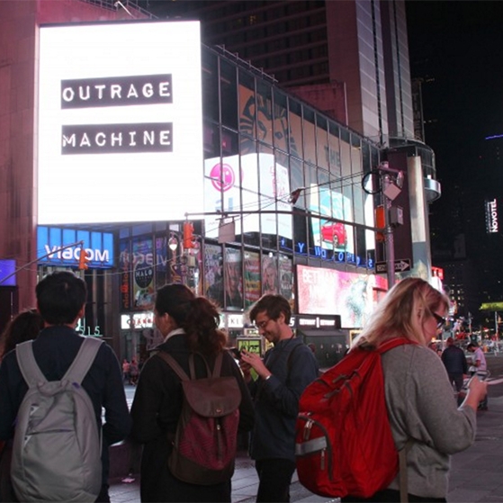 Times Square with the logo of Outrage Machine on display, with a few people looking at their cell phones