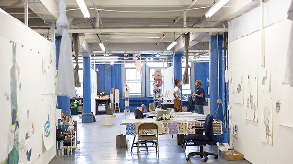 2 people talking in a warehouse space with a lot of drawings on the wall and designs spread out on a table.