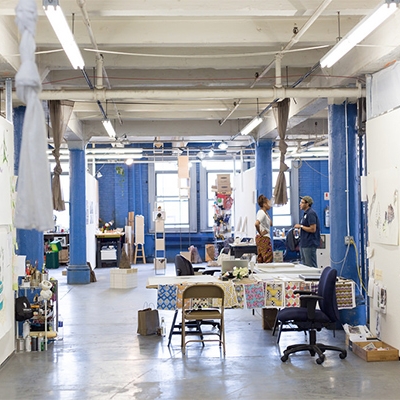 2 people talking in a warehouse space with a lot of drawings on the wall and designs spread out on a table.