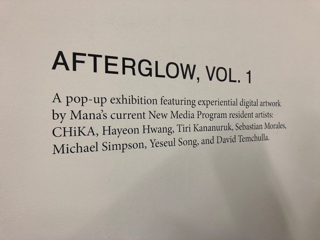 A Plaque that reads "Afterglow Vol.1, A pop-up exhibition featuring experiential digital artwork by Mana's current New Media Program resident artists: CHiKA, Hayeon Hwang, Tiri Kananuruk, Sebastian Morales, Michael Simpson, Yeseul Song, and David Temchulla