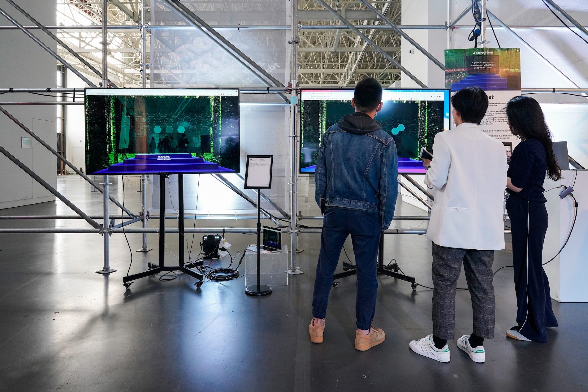 Two young men and a young woman stand with their backs to the camera as they watch two large screens display computer generated imagery