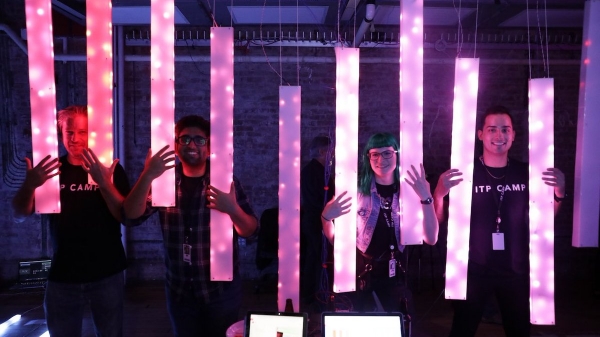 Four people wearing ITP Camp t-shirts stand smiling in a darkened room behind an installation of multiple, long, rectangular lights.