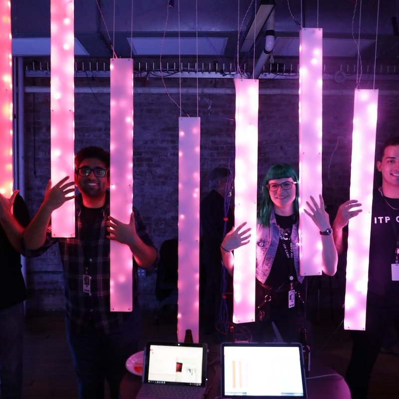 Four people wearing ITP Camp t-shirts stand smiling in a darkened room behind an installation of multiple, long, rectangular lights.