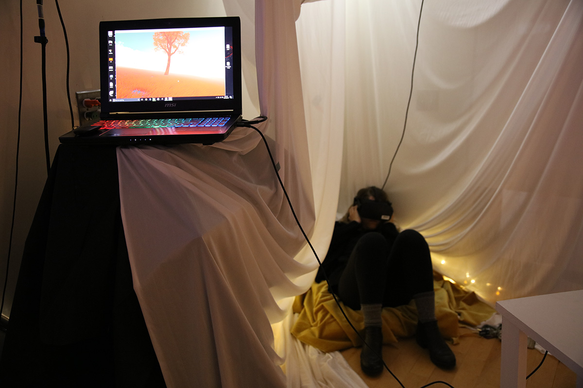 A person sitting on the floor wearing VR googles with a monitor showing a solitary tree in open space