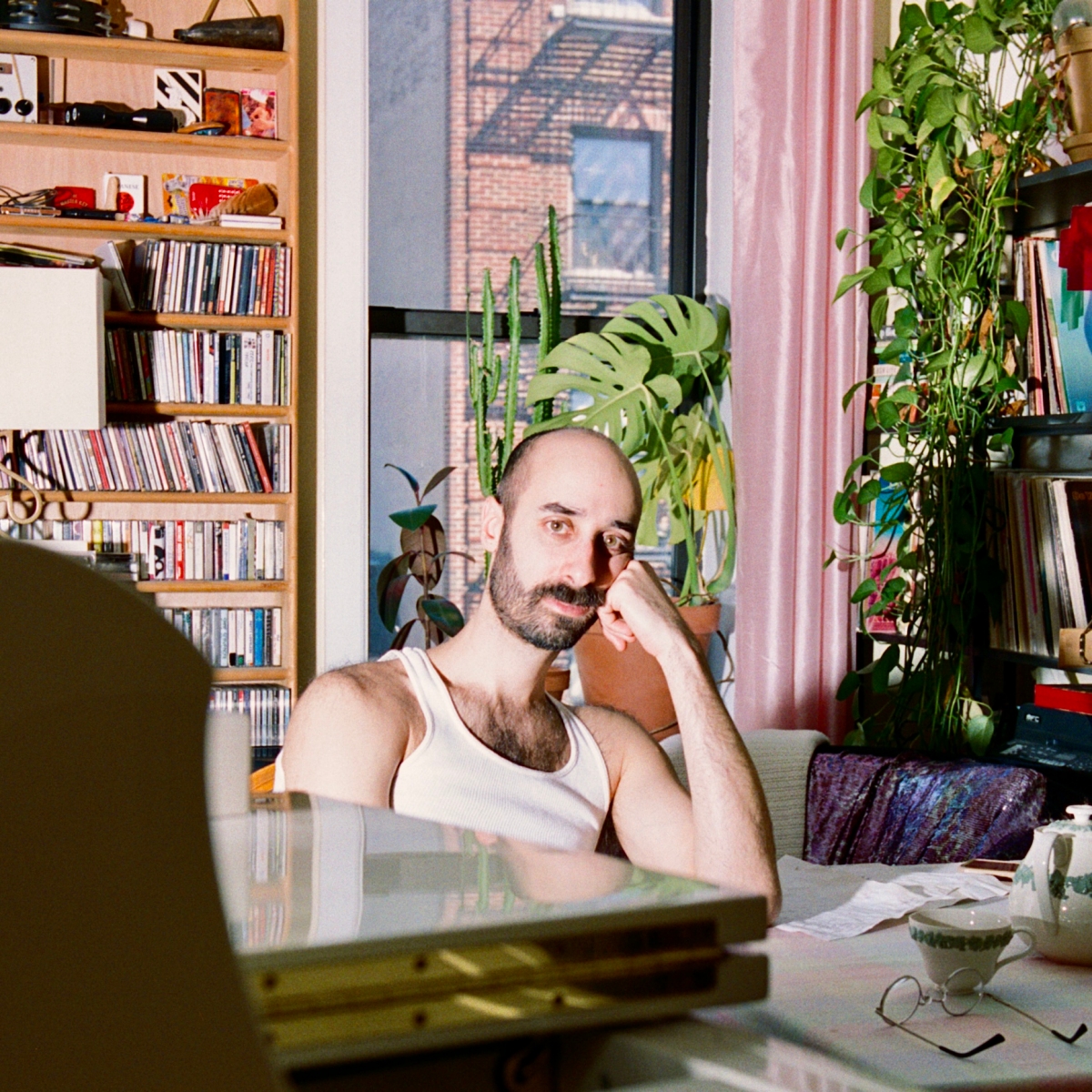 Ron Shalom leans against a desk, with bookshelf, window, and plants in the background