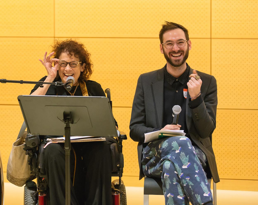 On the left, a white woman in a wheelchair with curly hair adjusts her glasses, smiling. On the right, a bearded white man laughs, mid-snap.