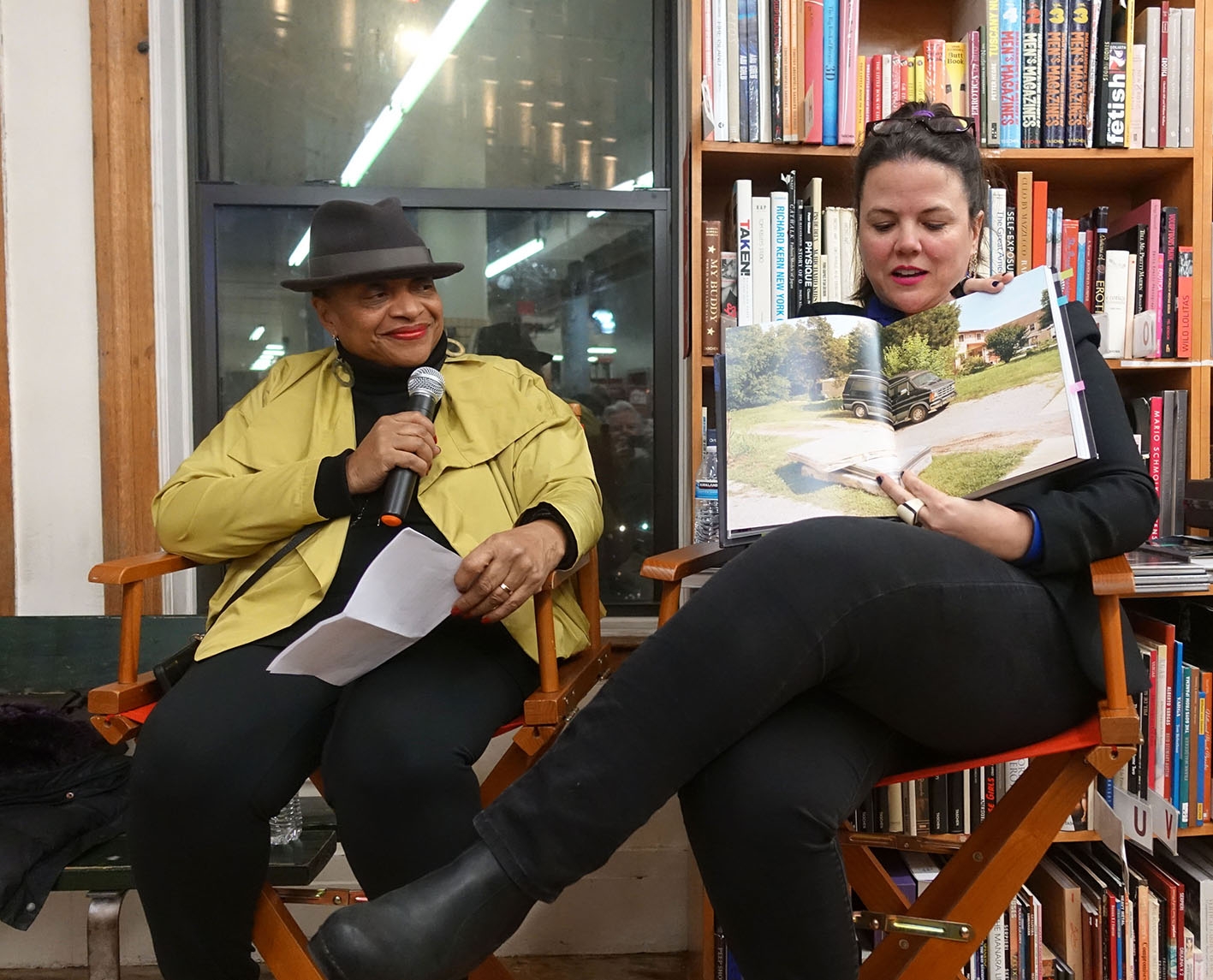 Two women sitting in chairs in a bookstore. One holds a book open to talking about the photos within, the other holds a mic and look on.