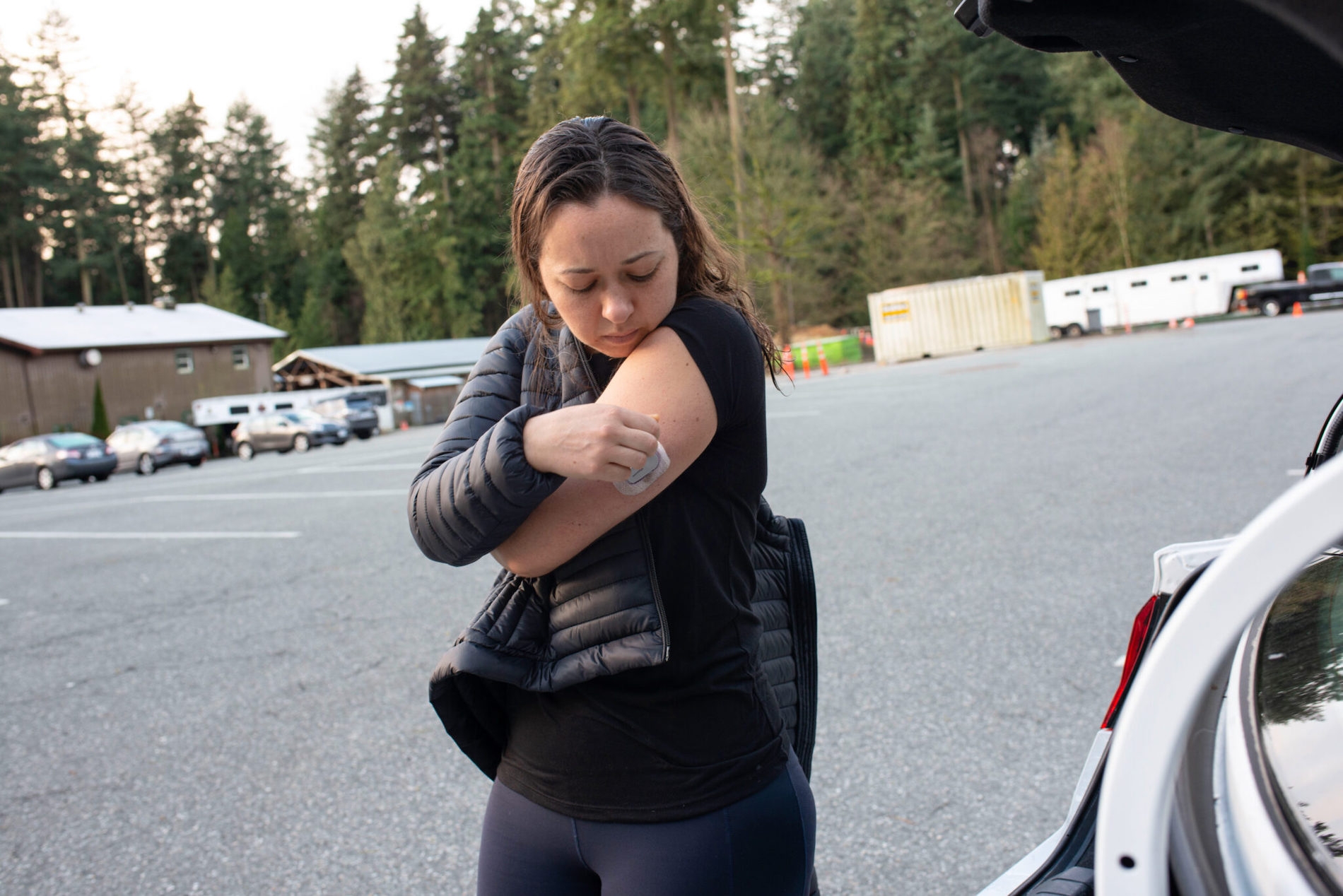 A woman in black puts a patch on her arm in a parking lot surrounded by trees.