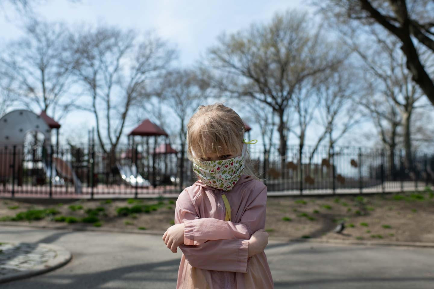 A little girl with a face mask on crosses her arms in front of gated playground