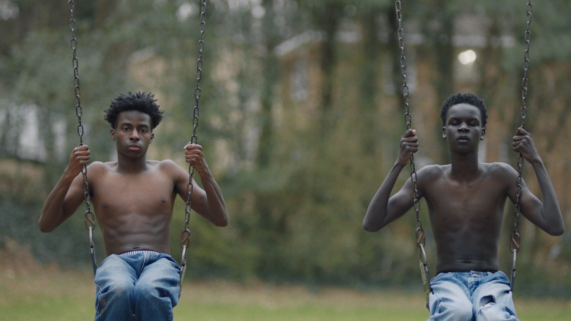 Two black teenage boys sit side by side on swings facing the camera, shirtless, wearing blue jeans.