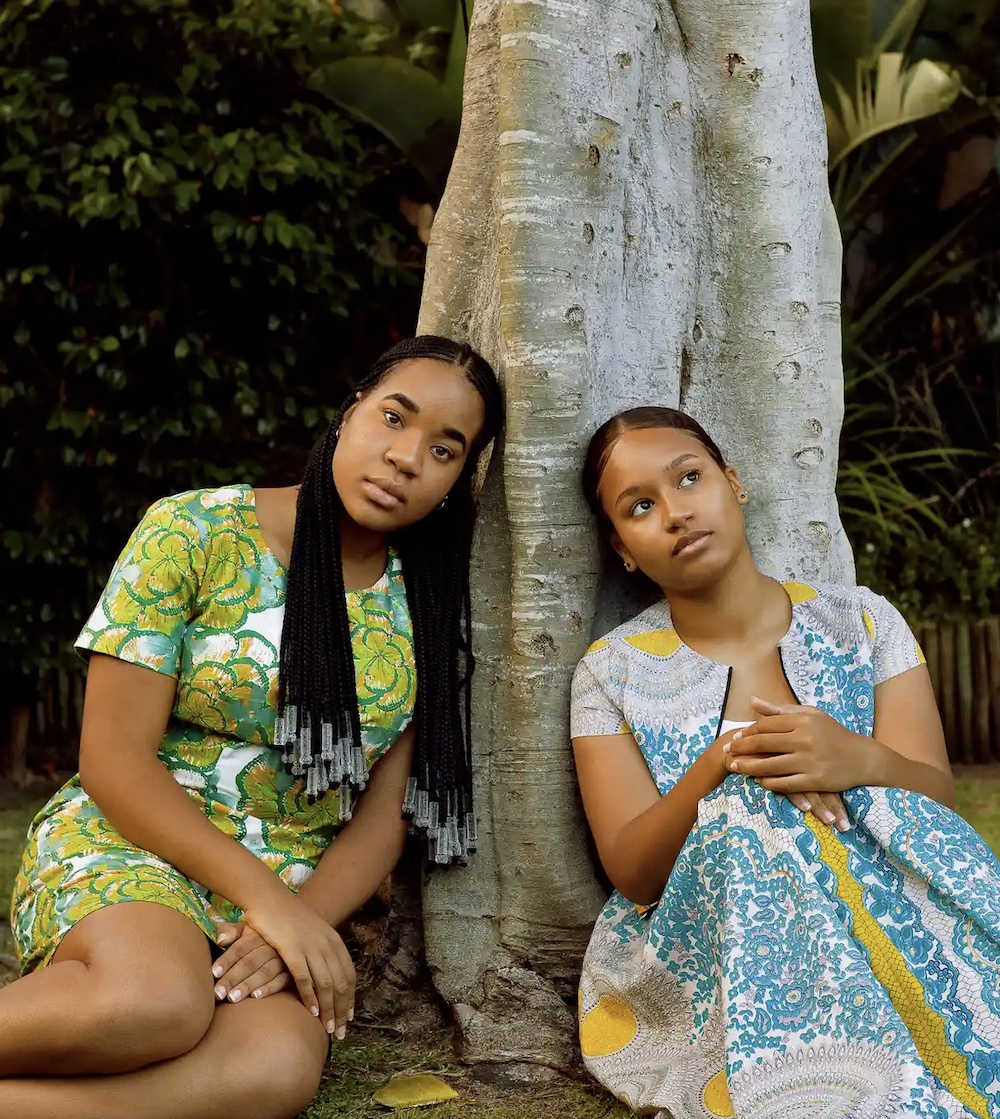 two women sit at the foot of a tree, both wear a colorful dress and one looks onward at the camera while the second gazes upward