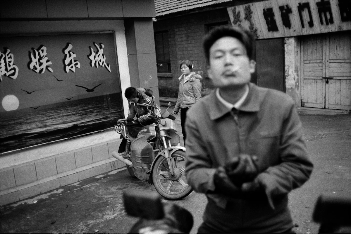 a man faces the camera smoking a cigarette while a couple in background sits on a motorcycle