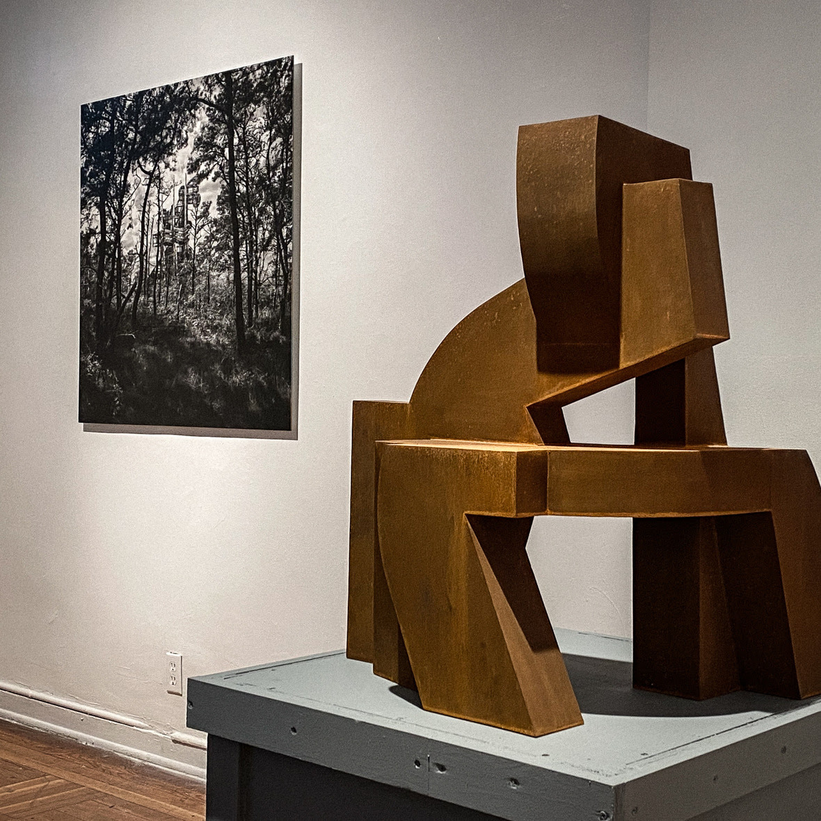 an interior gallery view contains an abstract sculpture set before a wall with a square two dimensional work of art