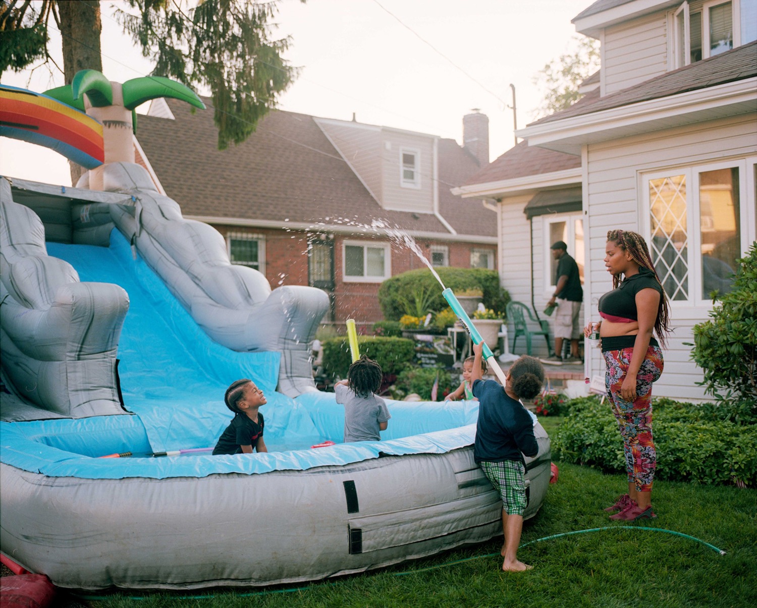 a family stands outdoors in yard playing with water toys and an inflatable water slide and pool