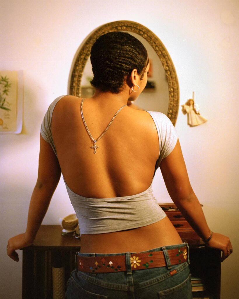 A Black woman looks at herself in the mirror with her back to the camera. 