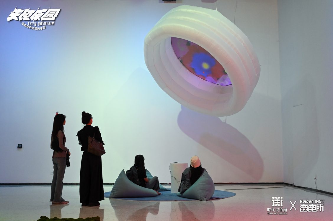 Two people stand to the left observing two museum-goers seated on the floor interacting with an inflatable pool mounted to the ceiling