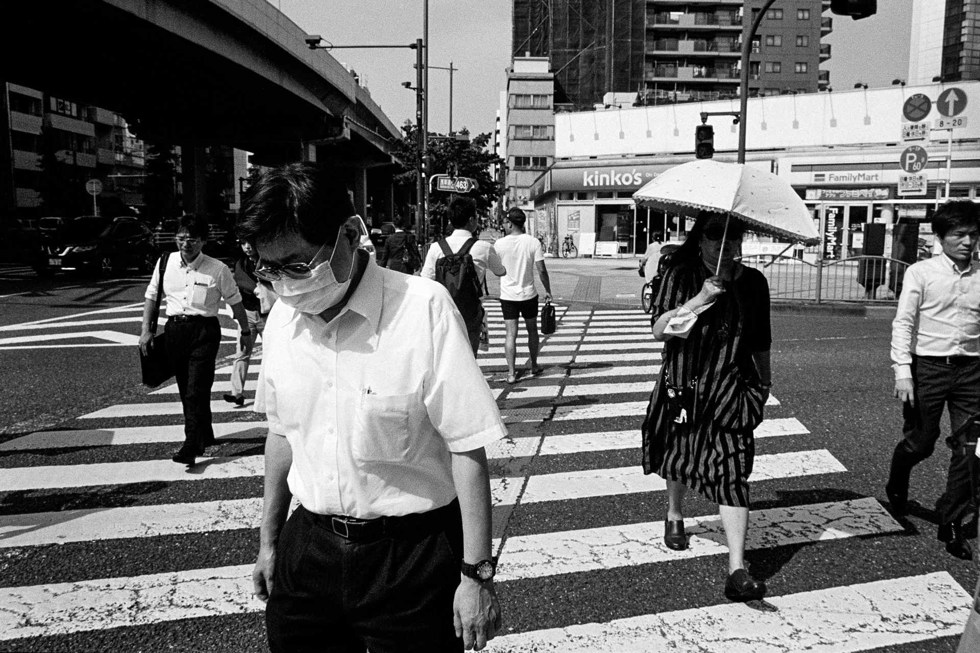 black and white photo of people crossing street, man in foreground wearing a medical mask