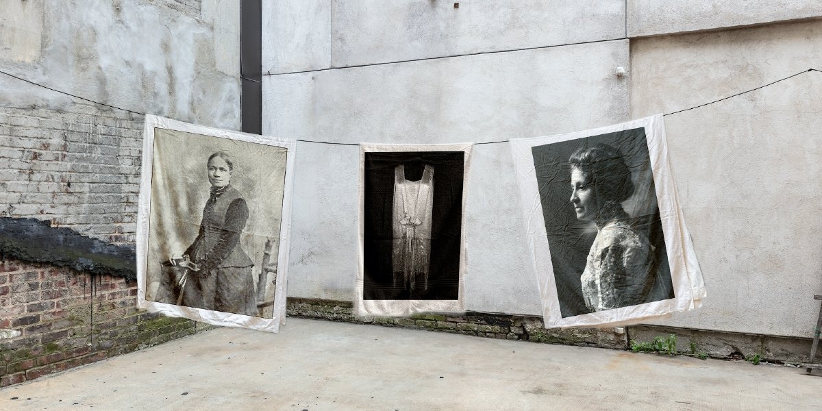 three tapestry style black and white portraits hand on clothes line in an exterior. Two portray a woman in profile, while a third displays a dress in posed placement