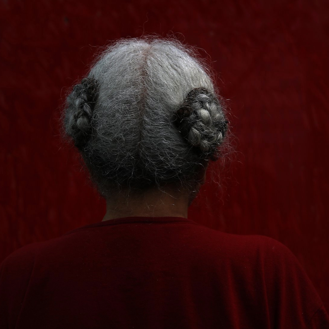Image of the back of an older woman with grey hair pulled into two buns standing in front of a red wall. 
