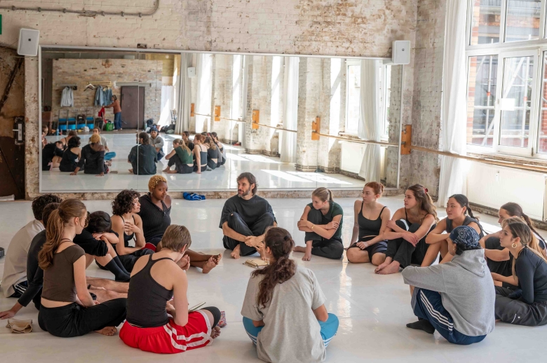 A group of dancers sitting in a circle during a training session in a dance studio