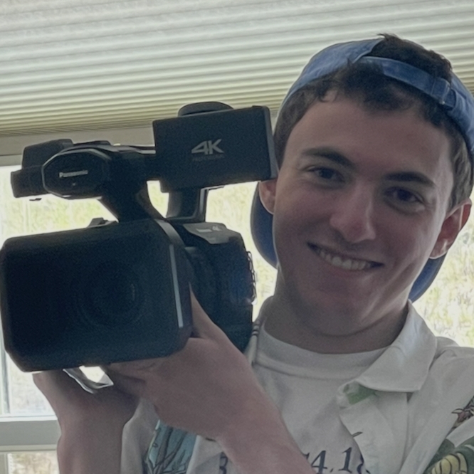 Student wearing a hat and holding a film camera on his shoulder.
