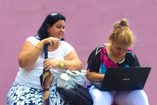 Two women seated, one looking over the other's shoulder as she types on a laptop computer.