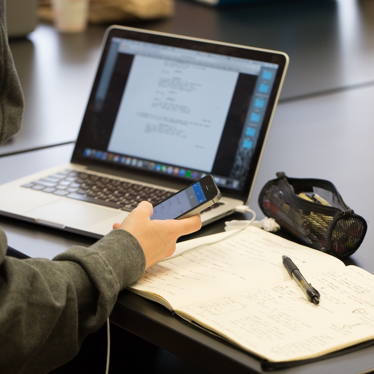 Student holding a phone and looking at a script open on a laptop computer.