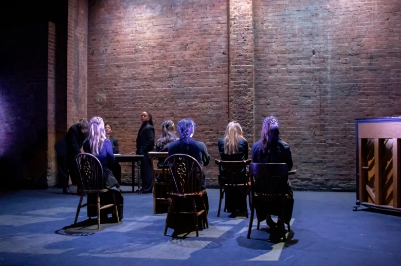 Actresses sitting in chairs facing the wall with one actress still standing and another leaning over a table during a scene from the Spring 2026 final presentation of "Measure for Measure".