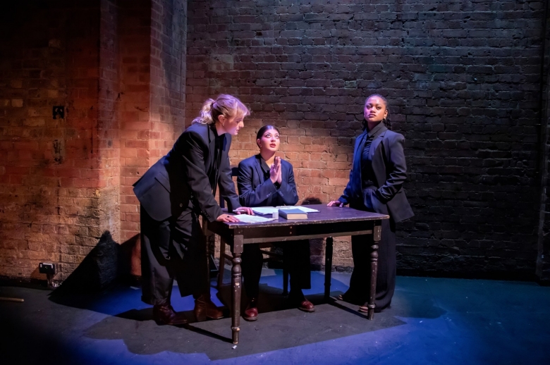 Actresses gathering at a table. One is sitting with prayer hands looking towards the sky. The other two are standing near the table during a scene from the Spring 2026 final presentation of "Measure for Measure".