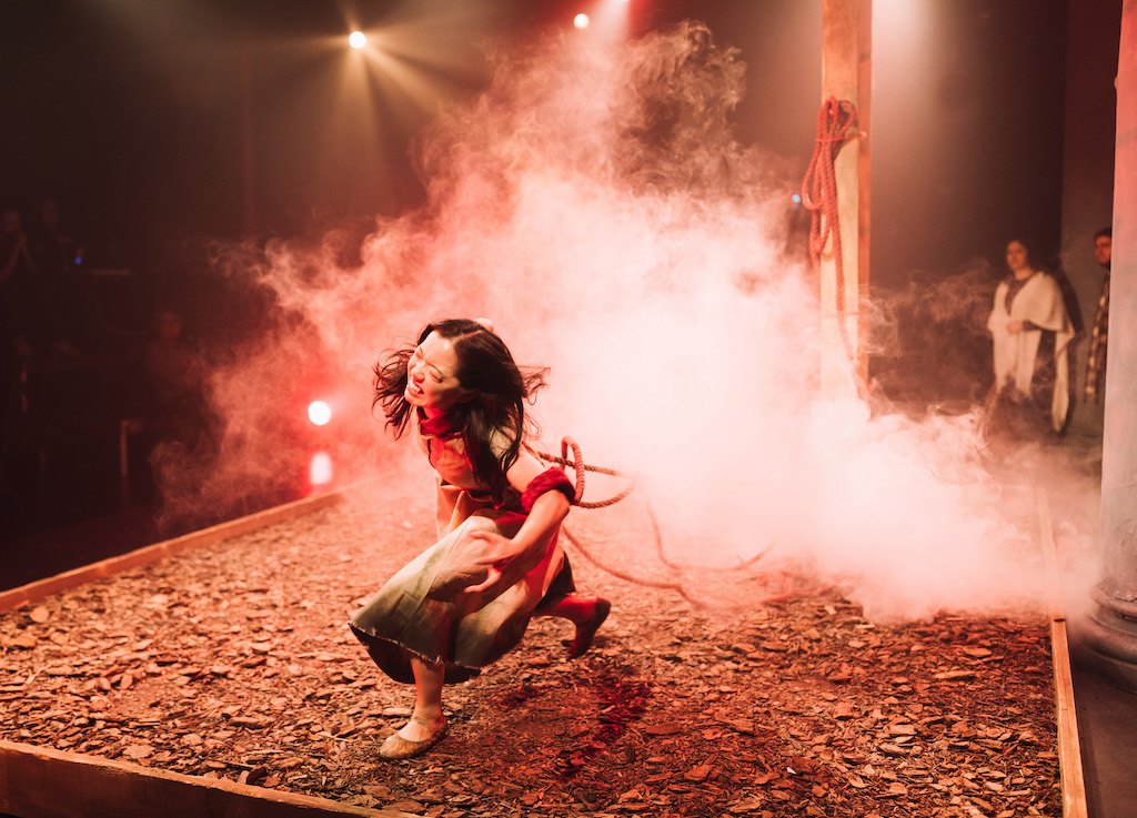 Actress lunging forward on stage, mid-performance, lit with red light and theatrical smoke