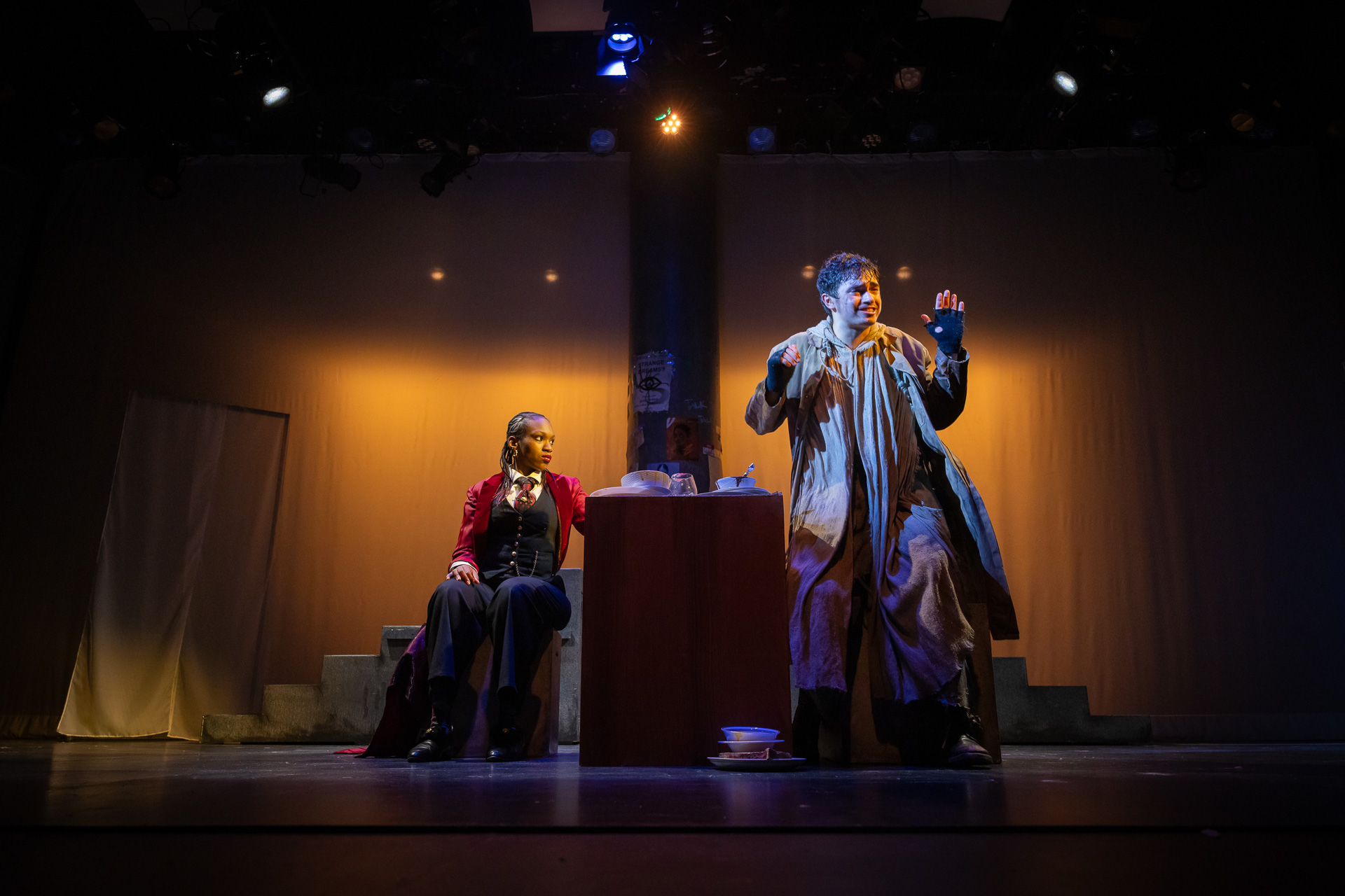 two people on a dimly lit stage, one seated, the other standing up and speaking