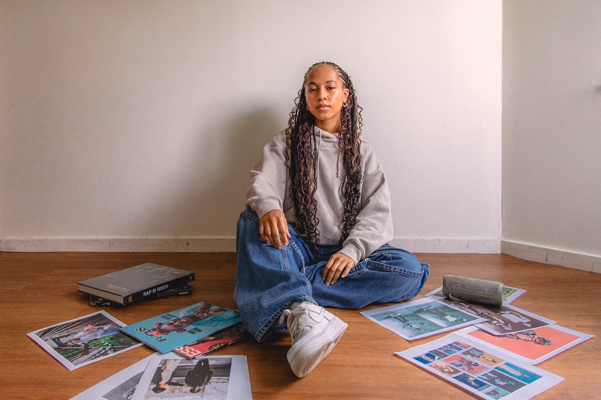 Denise Stephanie Hewitt sits against a wall on a hardwood floor with photos surrounding the floor around her