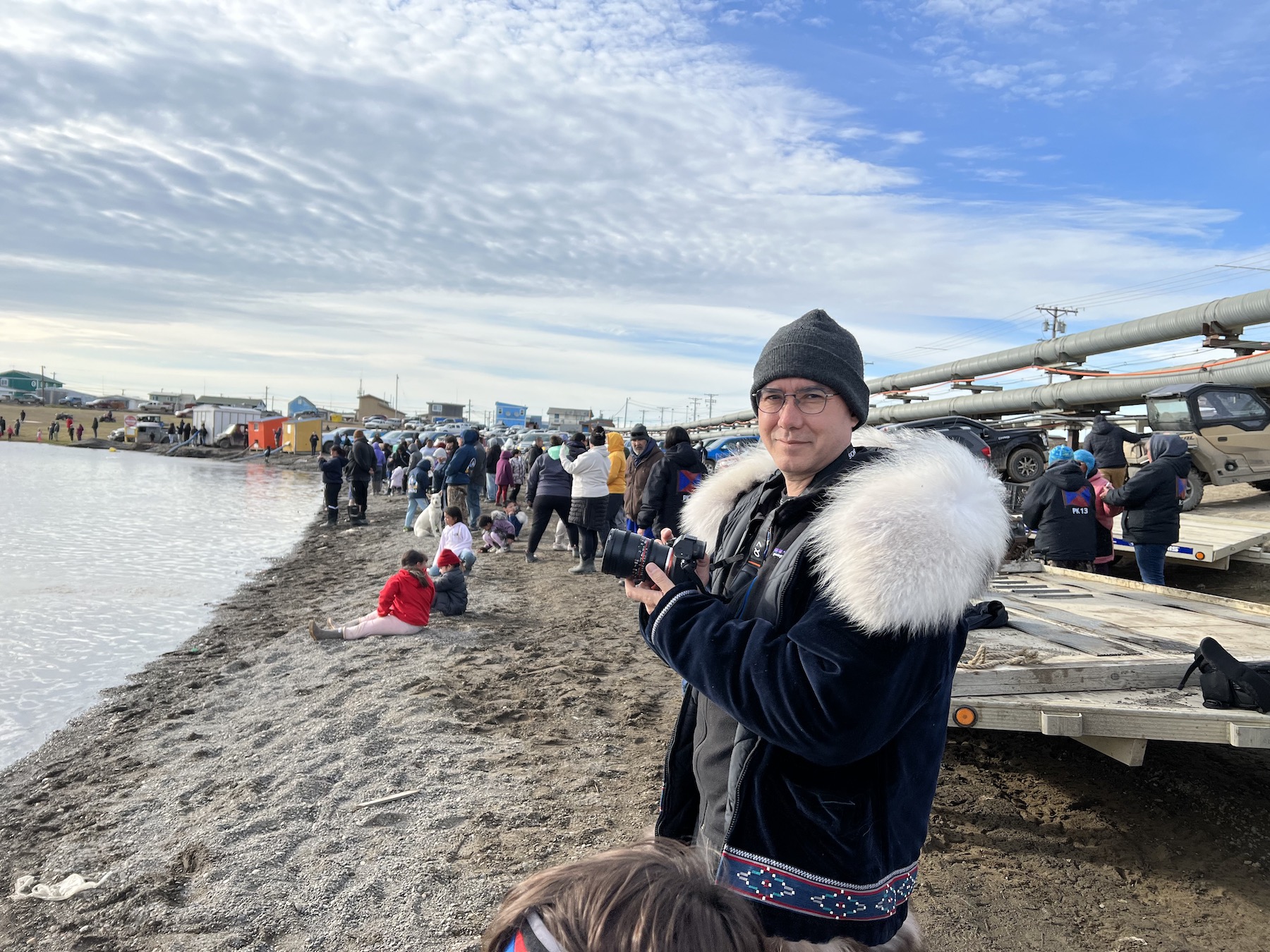 MacLean stands in a fur coat on a beach holding a camera